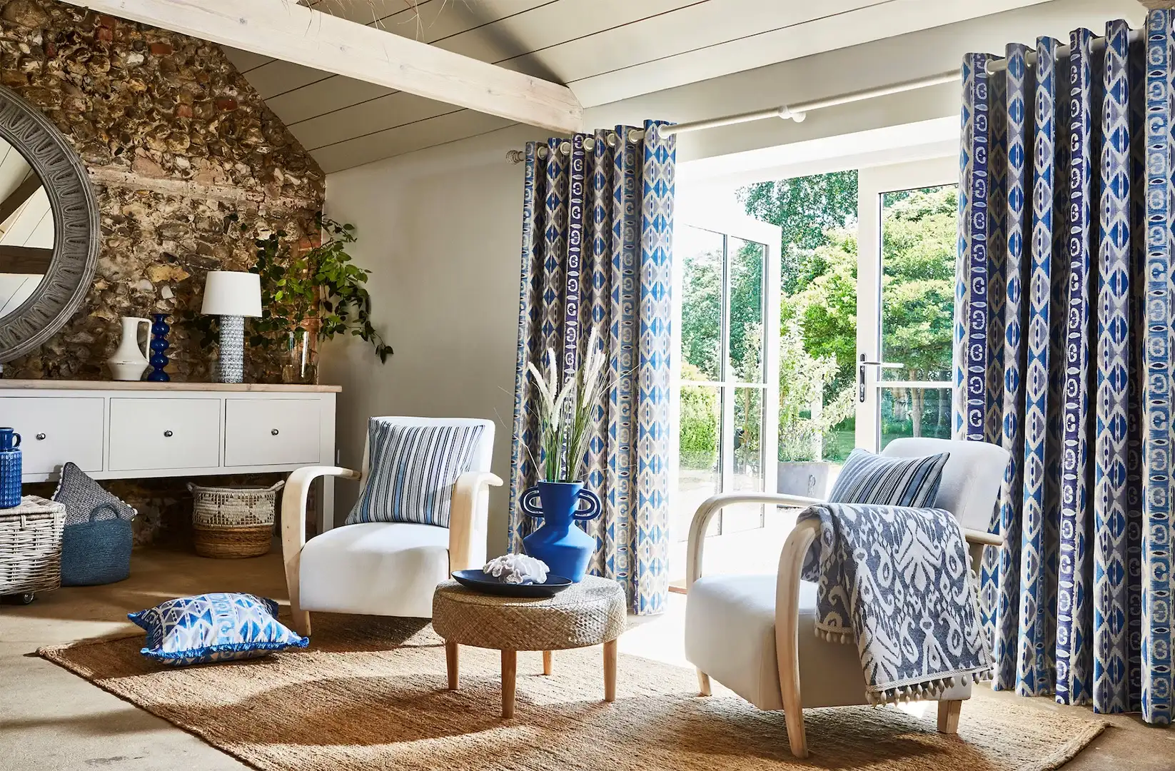 A bright sitting room with blue and white patterned curtains framing open garden doors